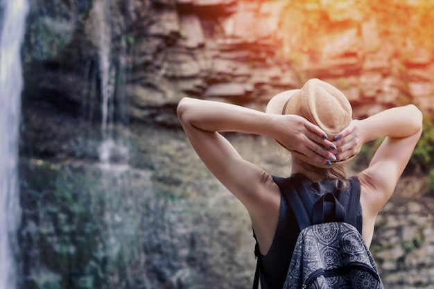 Chica con un sombrero con mochila mirando una cascada.