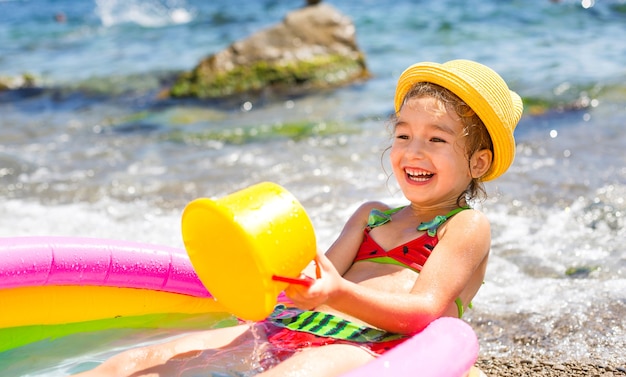 Chica con sombrero de paja amarillo juega con el viento, el agua y un dispensador de agua en una piscina inflable en la playa.