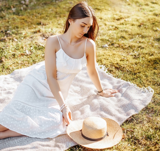 Chica con sombrero de paja en la primavera en el parque. Morena de pelo largo sentada sobre un plaid sobre un fondo de naturaleza veraniega. Juventud y belleza.