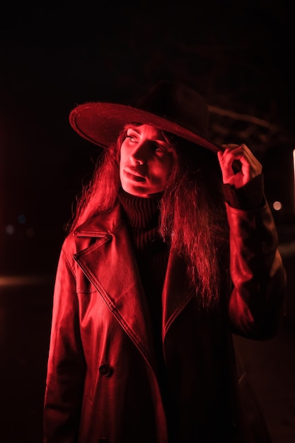 Chica con un sombrero en un retrato por la noche con un impermeable en un paquete. Iluminado con luz roja tenue desde el lateral