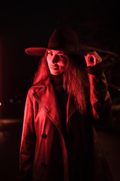 Chica con un sombrero en un retrato por la noche con un impermeable en un paquete. Iluminado con luz roja tenue desde el lateral