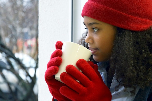 Chica con sombrero rojo y guantes con una taza mirando por la ventana