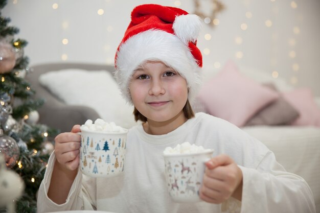 Chica con sombrero de Santa Claus bebe cacao con malvaviscos en medio del árbol de Navidad