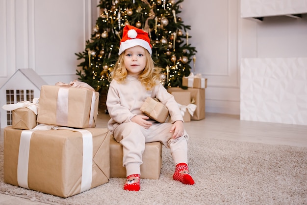Chica con sombrero de Santa con regalo de Navidad sobre fondo de árbol de Navidad. Niño con regalo de Navidad en casa. Casa decorada para las vacaciones de invierno. Enfoque suave