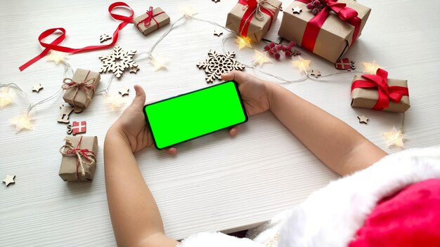 Chica con sombrero de Santa con smartphone con pantalla verde sobre fondo blanco de mesa de madera con regalos de Navidad.