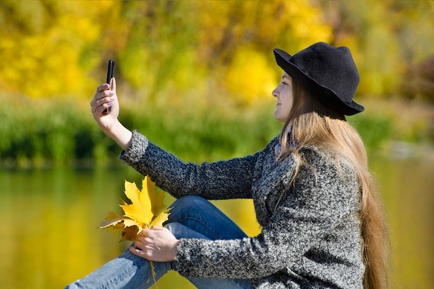 Chica del sombrero sentada en el muelle y hace selfie