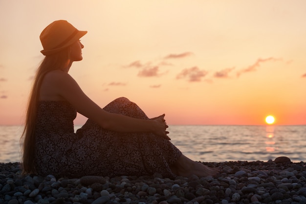 Foto chica con un sombrero sentado en el mar al atardecer.