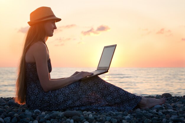 Chica con un sombrero sentado y trabajando en su computadora portátil contra el mar al atardecer