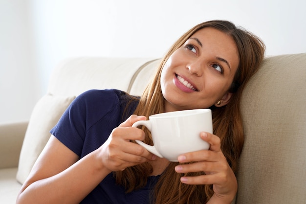 Chica soñadora bastante positiva sostiene la taza con bebida caliente imaginando sentado en el sofá