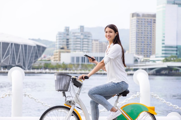 Chica sonriente en auriculares sosteniendo teléfono inteligente y montando bicicleta en el parque