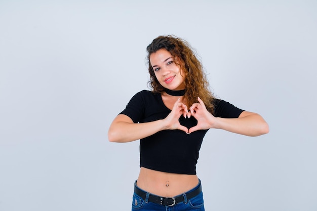 Chica sonriente está mostrando gesto de corazón sobre fondo blanco.