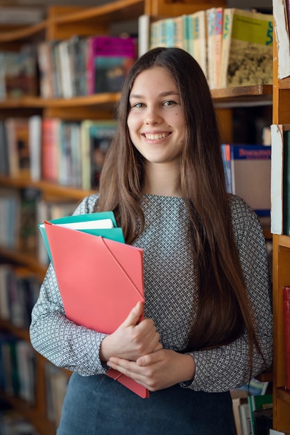 Chica sonriente feliz con carpetas en la biblioteca Chica estudiante diligente ansiosa por estudiar y ampliar su conocimiento