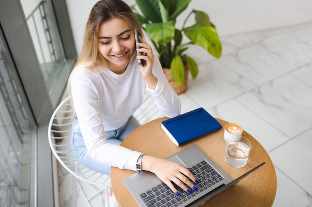Chica sonriente hablando por teléfono móvil mientras está sentada en una cafetería durante y trabajando en una computadora portátil