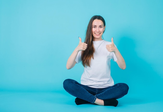 La chica sonriente muestra gestos perfectos al sentarse en el suelo con fondo azul.