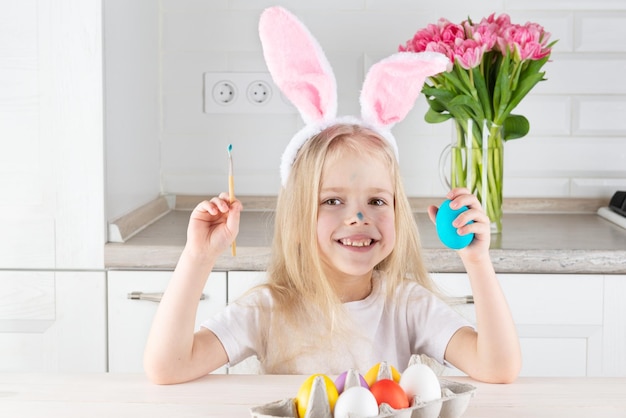 Chica sonriente con orejas de conejo con un cepillo y huevos de Pascua en la mesa