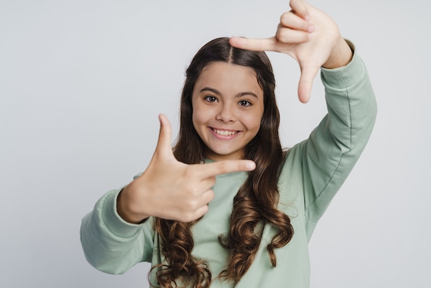 Chica sonriente y positiva posando sobre un fondo de pared blanca. El niño muestra un marco con sus dedos.