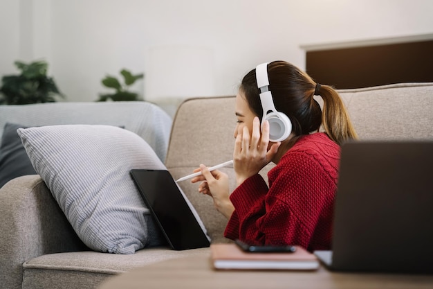 Chica sonriente relajándose en casa, está tocando música usando una computadora portátil con tableta de teléfono inteligente y usando auriculares blancos