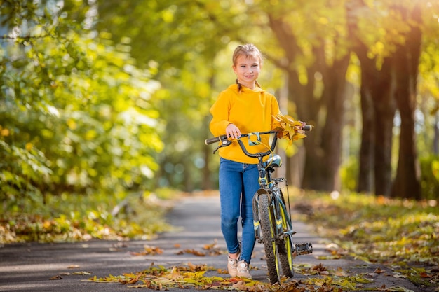 Chica sonriente en suéter amarillo camina con bicicleta en un parque de otoño en un día soleado y agita una mano amistosa