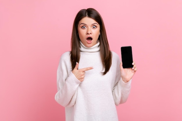 Chica sorprendida con la boca abierta apuntando a la pantalla vacía del teléfono inteligente con espacio de copia para publicidad usando un suéter blanco de estilo casual Foto de estudio interior aislada en fondo rosa