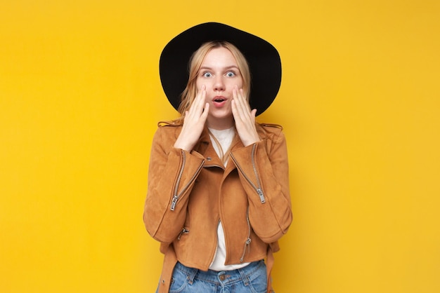 Una chica sorprendida con una chaqueta y un sombrero de otoño se sorprende con un fondo amarillo aislado