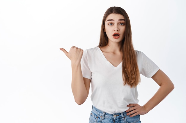 Chica sorprendida y curiosa apuntando a la izquierda a algo interesante, haciendo preguntas sobre el producto, haciendo preguntas, interesada con el logo de la pancarta, de pie sobre una pared blanca