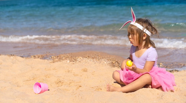 Chica sorprendida con diadema de conejito en una falda rosa sentada con huevos coloridos en la playa de arena