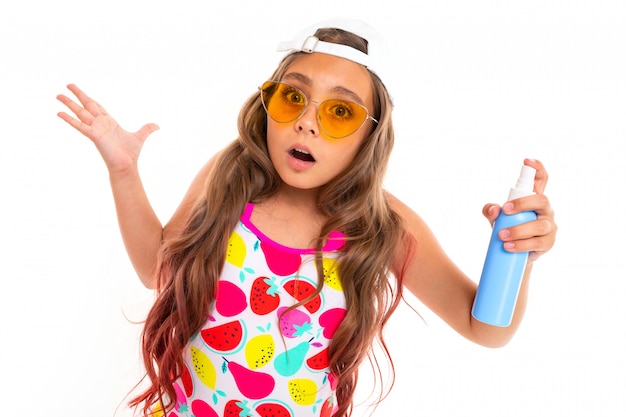 Chica sorprendida en un look de verano sostiene una botella de protector solar en una pared blanca