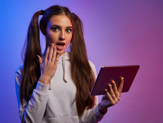 Chica sorprendida con tablet pc digital, fondo de estudio de neón. Mujer joven sorprendida con almohadilla, boca cerrada con la mano.