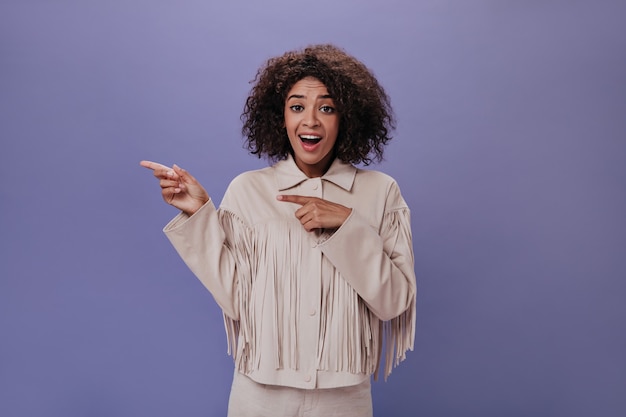 Chica sorprendida en traje beige con flecos muestra su dedo a un lado