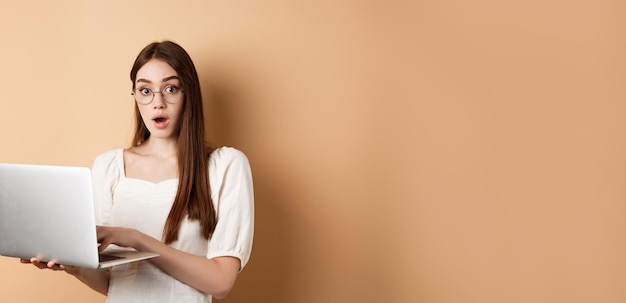 Chica sorprendida usando una computadora portátil y jadeando asombrada dice wow mirando con gafas a la cámara de fondo beige