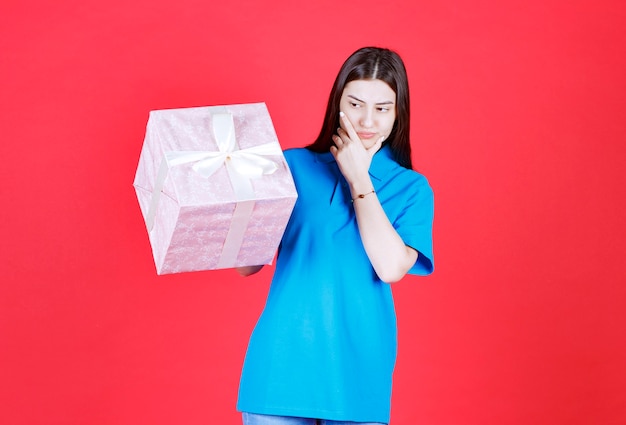 Chica sosteniendo una caja de regalo púrpura envuelta con cinta blanca y parece confundida y pensativa.