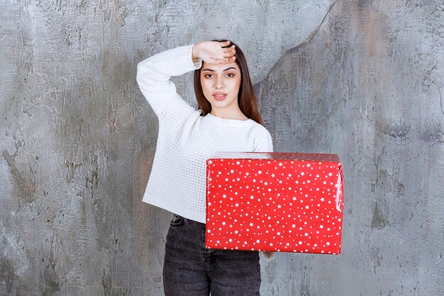 Chica sosteniendo una caja de regalo roja con puntos blancos y parece cansada.