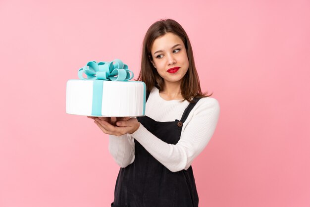 Chica sosteniendo un gran pastel aislado en pared rosa con expresión triste