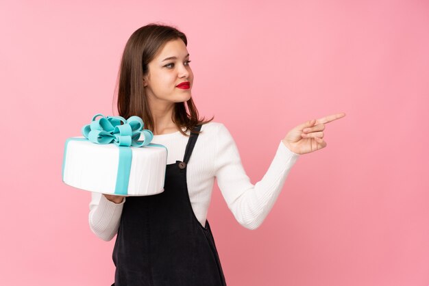 Chica sosteniendo un gran pastel en la pared rosa apuntando hacia el lado