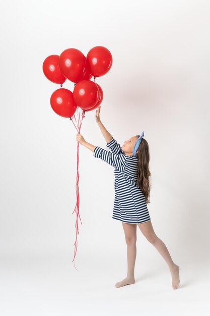 Chica sosteniendo en la mano globos rojos, la otra mano alcanza globos y de puntillas en blanco