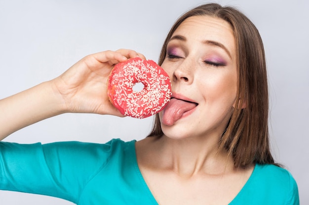 Chica sosteniendo y mostrando rosquilla rosa tratando de chupar con la lengua fuera en fondo gris