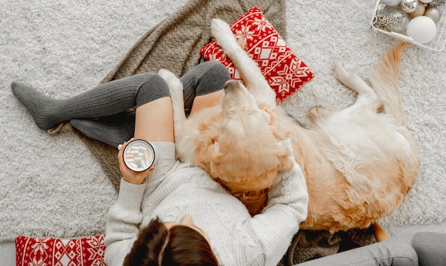 Chica sosteniendo una taza con cacao y malvavisco y abrazando a un perro golden retriever en las vacaciones festivas. Mujer joven con bebida caliente y perrito mascota en casa