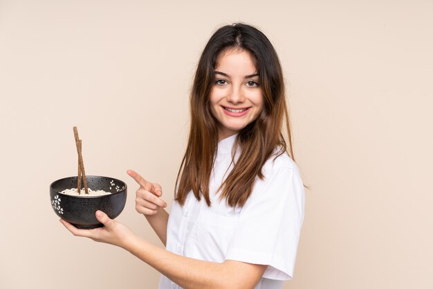 Chica sosteniendo un tazón lleno de fideos sobre pared aislada y apuntando