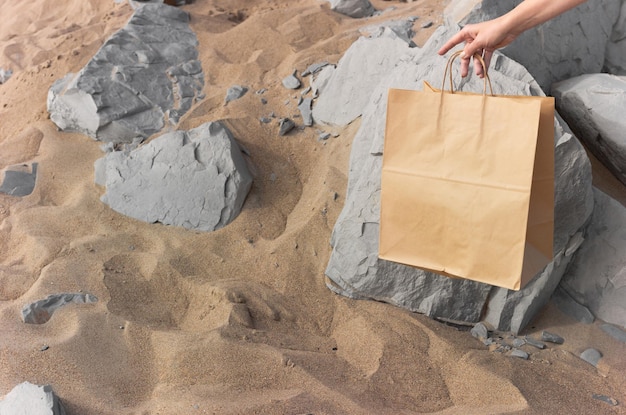 Foto chica sostiene una bolsa de papel artesanal beige en el fondo de una playa con arena y rocas