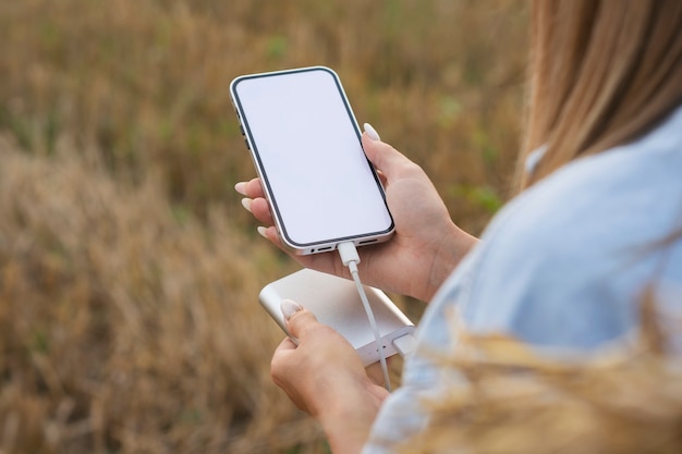 Chica sostiene una maqueta de un teléfono inteligente con una pantalla blanca en sus manos. Power Bank carga el teléfono en el contexto de la naturaleza.