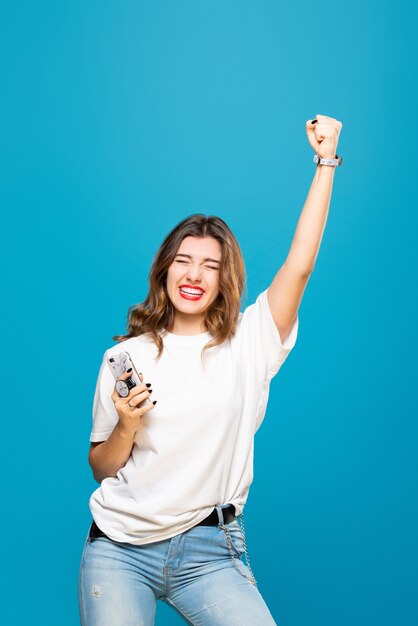 Foto chica sostiene el teléfono en sus manos con una sonrisa en su rostro y levanta su mano sobre un azul