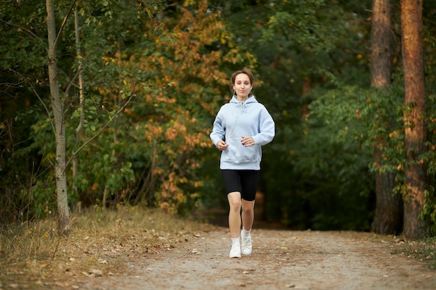 chica con una sudadera azul y auriculares en el parque y corriendo. chica trotando en el parque.