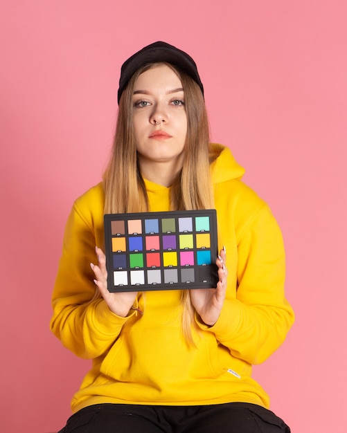 Una chica con una sudadera con capucha amarilla y una gorra negra está en el estudio sosteniendo un corrector de color en sus manos, el fondo detrás de ella es rosa.