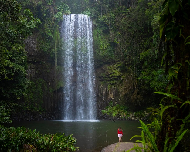 Una chica con una sudadera roja y una falda roja y blanca se alza sobre una poderosa cascada tropical en aus