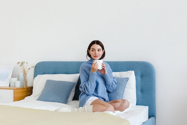 Chica en un suéter azul en el interior de estilo Hygge con una taza de té caliente en sus manos se sienta en la cama