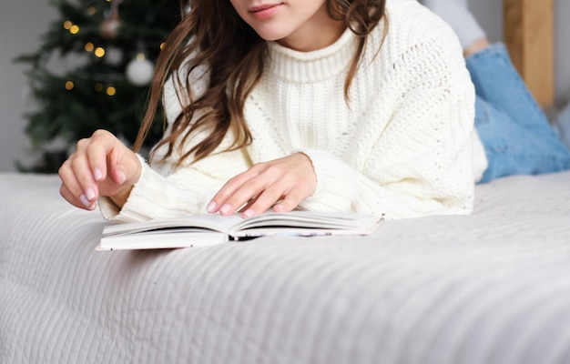 Una chica con un suéter blanco se acuesta en la cama y lee un libro.