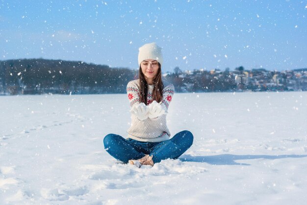 Chica en un suéter blanco de invierno Se sienta con las piernas cruzadas en la nieve Lanza la nieve