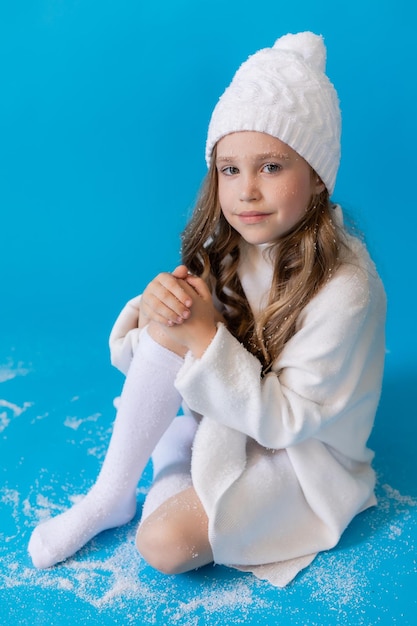 chica en un suéter blanco y sombrero sopla nieve artificial de la palma en el estudio sobre fondo azul