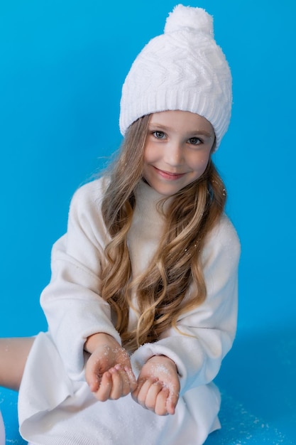 chica en un suéter blanco y sombrero sopla nieve artificial de la palma en el estudio sobre fondo azul