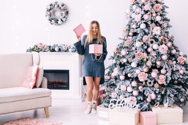 Chica con un suéter gris abrió la caja de regalo en una acogedora habitación con chimenea, sofá y árbol de Navidad.